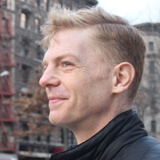 Christopher Moffett in profile, smiling, blonde short hair, red brick background