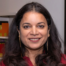 Manisha smiling at the camera, long dark and wavy hair, gold earings, red top