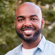 Omari smiling at the camera, shaved head, mustache and beard, blue shirt