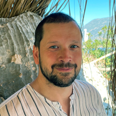 James Thurman smiling at the camera, short dark hair, beard, striped shirt, outdoor background