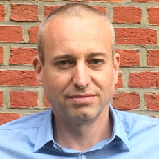 Joris looking at the camera, short hair, blue shirt, red brick background