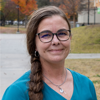 Kim Willis facing forward, smiling, hair in a braid, wearing glasses and a blue top.