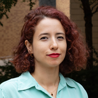 Nasrin is facing forward. She has red, curly hair, bright red lipstick and wears a turquoise shirt.