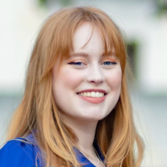 Caitlin is facing forward and smiling. She has long red hair and wears a blue top.