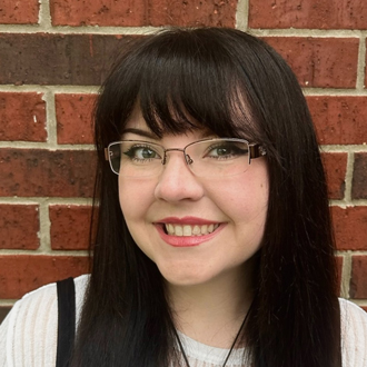 Raegan is facing forward and smiling. She has dark long hair with bangs and wears glasses.