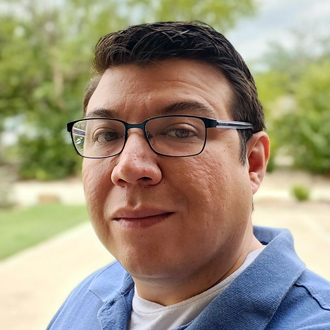 Andres is facing forward. He has dark hair, glasses and wears a blue shirt.