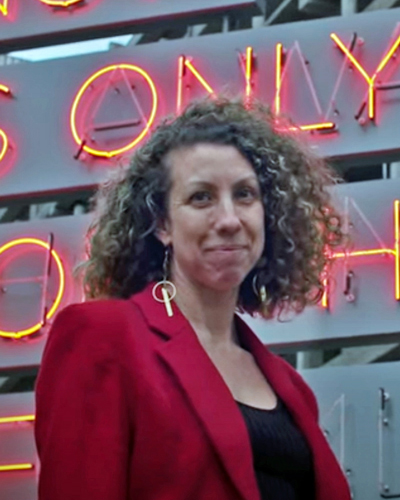 Alicia smiling at the camera, curly hair, red jacket, sign in the background.