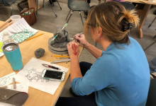 Image of ceramics workshop participant sitting at table and working on sculpture. Sketches of projects and clay supplies scattered across table.