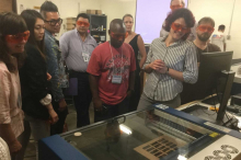 Image of a large group of workshop participants standing around equipment in the FabLab wearing protective eye equipment. Workshop facilitator shown providing information on the DigiFab process.